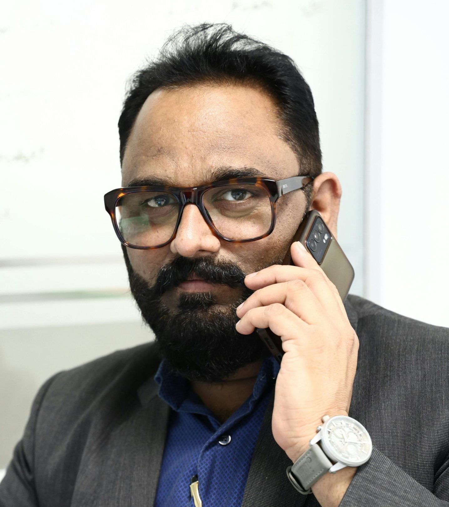 Confident businessman with beard and glasses speaking on mobile phone in office setting.